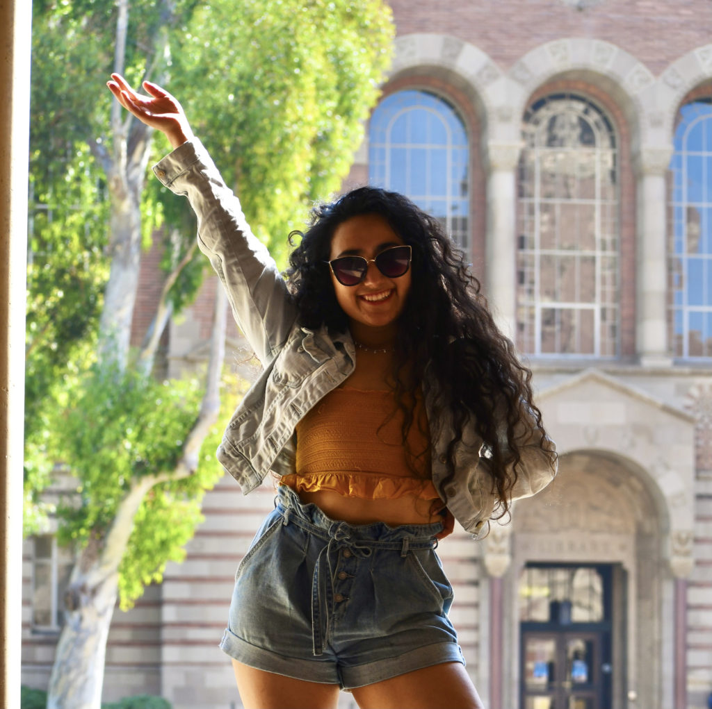 Nicole in jean shorts, yellow top, jean jackets and sun glasses waving in front of a brick building.