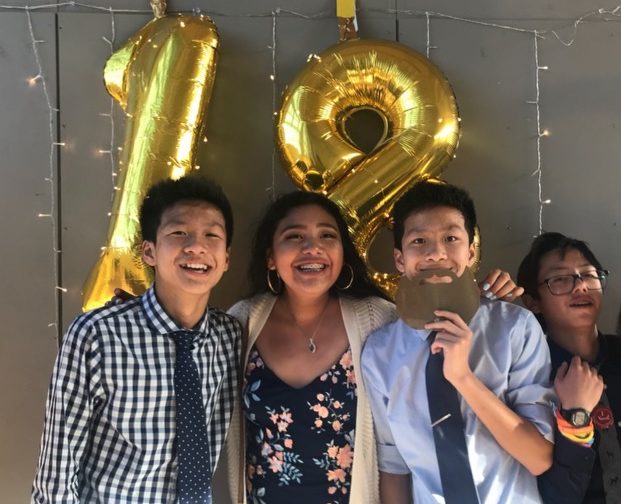 Cuatro jóvenes posando frente a un globo dorado con la forma del número 18.