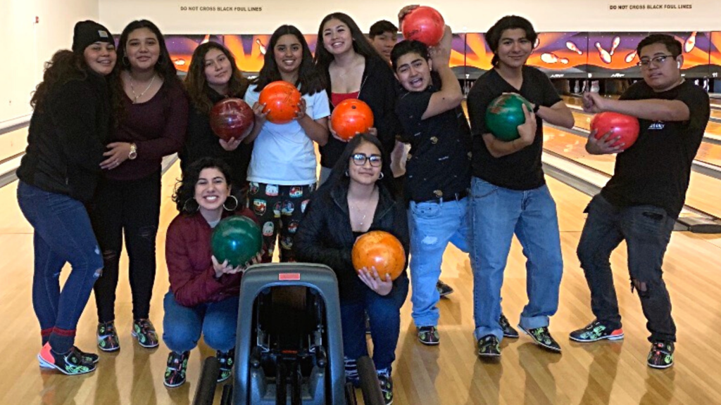 12 youth posing for the camera in front of a bowling lane with bowling balls in their hands.