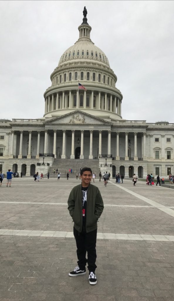 Juan in a brown jacket and black pants standing in front of the capitol building.