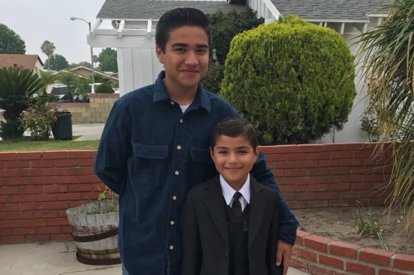 Juan in a blue button down shirt standing with a younger boy in front of a brick wall and houses.