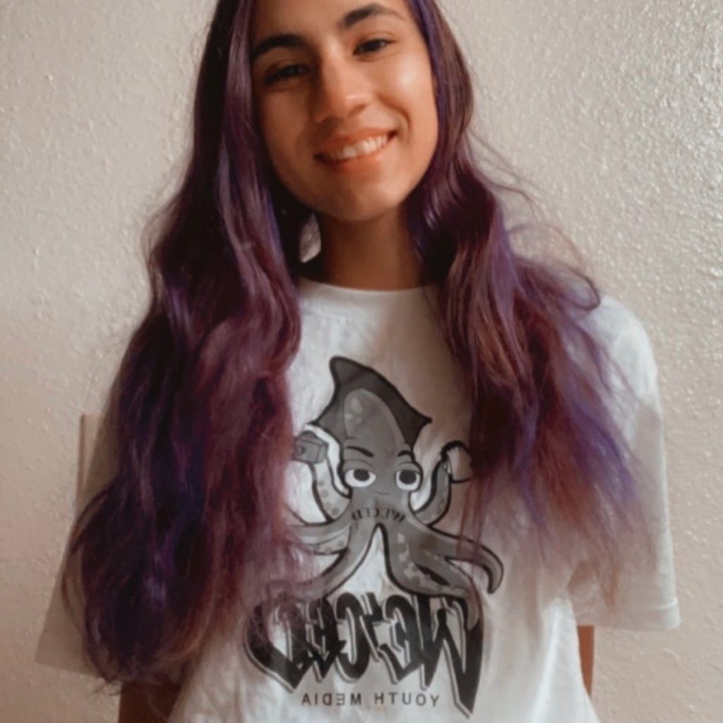 Stephanie is smiling with long purple hair and a white t-shirt with an octopus that says We'Ced