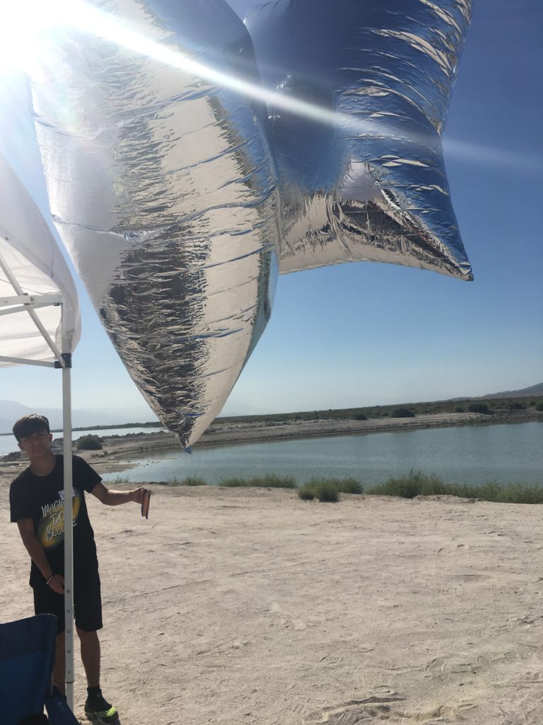 A young man in black t-shirt and beige shorts stands near a pop up tent. He is standing on a beach near the Salton Sea, and his holding two large silver balloons that are floating toward the camera.