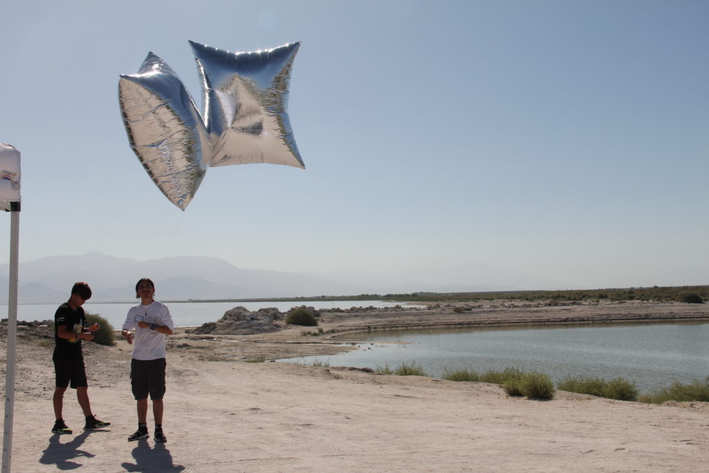 Two people standing on the end of the Salton Sea. They are holding strings attached to two large, square silver balloons.