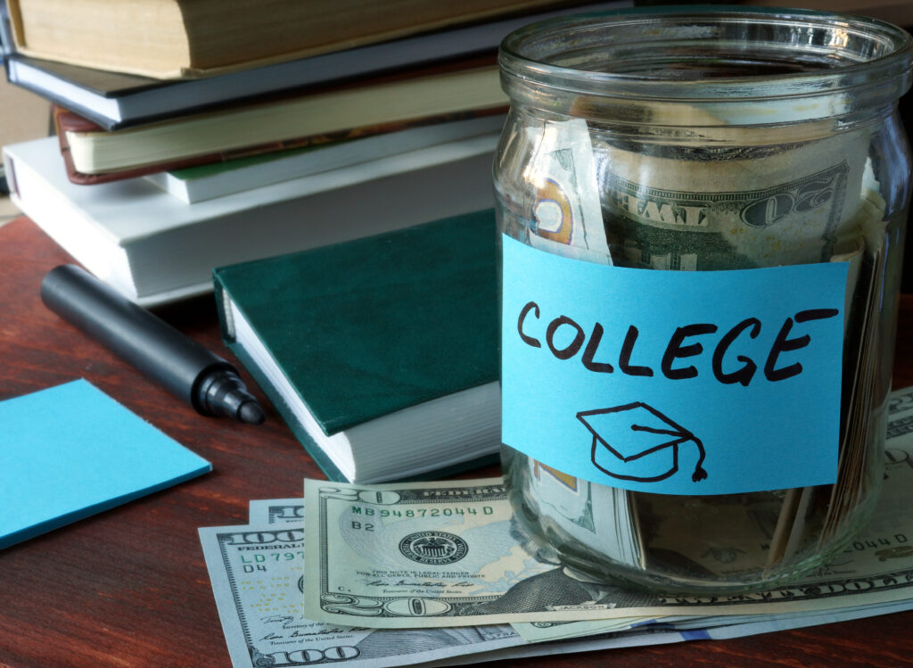 Mason jar full of 20 dollar bills with a tag reading "college" and a drawing of a graduation cap. The jar is sitting on more bills and has a stack of books behind it.